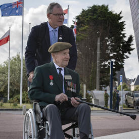 Un ancien combattant en fauteuil roulant, portant un béret et des médailles de guerre, est poussé par un homme. Plusieurs drapeaux sont en arrière-plan.