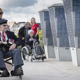 Two elderly people in wheelchairs, each pushed by a another person, pass in front of a monument.