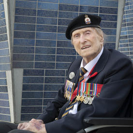 A veteran, wearing several medals is seated in a wheelchair, looking directly at the camera. A tall monument is behind him.