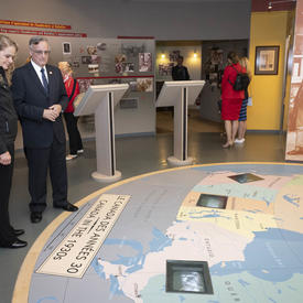 Governor General Julie Payette stands in a room while looking at a map on the floor in front of her. A man standing to her left is giving her explanations on what she is seeing.