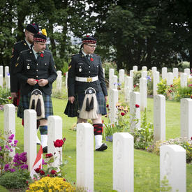 Three men in uniforms are walking in between two rows of tombstones in Bény-sur-Mer Canadian War Cemetery.