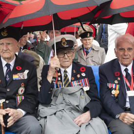 Three war veterans are seated in a front row of an audience with red and black umbrellas.
