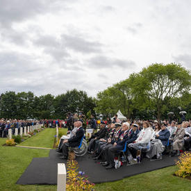 Une foule est assise lors d'une cérémonie au Cimetière de guerre canadien de Bény-sur-Mer.