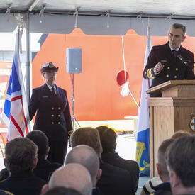 Jeffery Hutchinson, Commissioner of the Canadian Coast Guard, is speaking at a podium in front of a seated audience, under a white tent. A women wearing the Canadian Coast Guard Uniform is standing behind him, to his left, next to the Coast Guard flag.