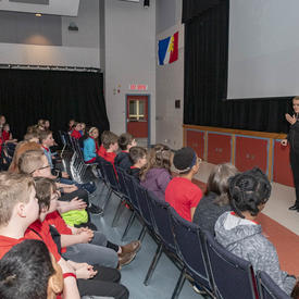 Un groupe d'élèves de la 1re à la 6e année sont assis sur des chaises devant la gouverneure générale Julie Payette, lors d'une présentation PowerPoint.