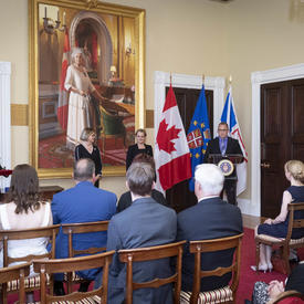 Governor General of Canada Julie Payette is standing in front of a small seated audience, while a gentleman is speaking at a podium  on her left. A large painting of Her Majesty Queen Elizabeth II is hanging behind her.