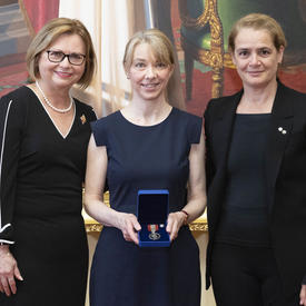 Une femme blonde tient une médaille dans une boîte de velours bleu entre Judy May Foote, lieutenante-gouverneure de Terre-Neuve-et-Labrador et Julie Payette, gouverneure générale.