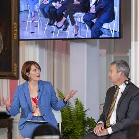 Alison Crawford, wearing a blue pant suit, is siting on a white tall stool, talking to David Aiken, wearing a grey suit and seated at a tall white stool. A large tv screen above them shows a panel of 5 people.