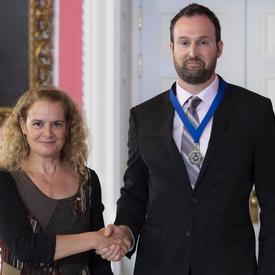 Governor General of Canada Julie Payette and reporter Greg Mercer, wearing a medal with a blue ribbon, are standing looking directly at the camera.