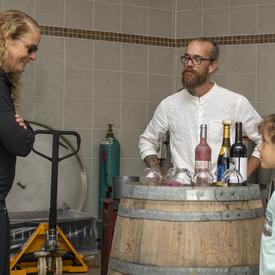 The Governor General speaks with a young girl and Laurence-Olivier Brassard inside the winery, where bottles of wine are displayed on a barrel.