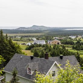 A photo of a white and grey manor at the Verger Poméloi hidden behind trees, land and mountains in the distance.