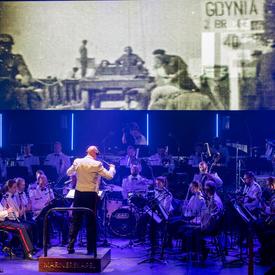 A photo of an orchestra on stage at the commemorative ceremony in the Scheldetheatre in Terneuzen, with footage of the Second World War playing on a large screen.