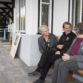 The Governor General is sitting with two women outside of the Cranberry Portage Heritage Museum.