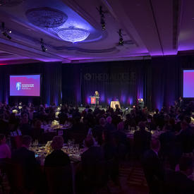 Governor General of Canada Julie Payette at a podium on stage, seen from the back of a large dark room with a few purple and pink lights. 