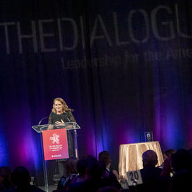 Governor General of Canada Julie Payette at a podium on stage. The stage is dark behind her. A few purple and pink lights are projected on a large black curtain behind her.