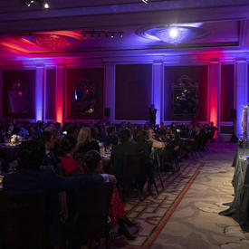 Side view of a room where Governor General Julie Payette is seen, at the far right of the photo, on stage, at a podium. The room is very dark but there are some purple and pink lights.