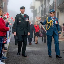 Governor General Julie Payette, wearing the Royal Canadian Air Force uniform, is waving to children holding small Canadian flags. The children are standing on the sidewalk.