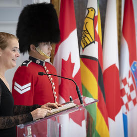 La gouverneure générale prononce une allocution à un podium, des drapeaux nationaux et un garde de cérémonie derrière.
