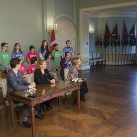 The Prime Minister, the Governor General and David Saint-Jacques`s wife, Véronique Morin, are sitting around a table looking at a television screen. A group of students are behind them. 