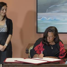 A lady sitting at a table is signing a document while a lady watches over her right shoulder.