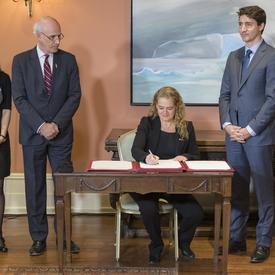 Governor General Julie Payette is sitting at a table, signing a document while a lady, the Privy Council Clerk Michael Wernick and Prime Minister of Canada Justin Trudeau watch her, standing. 
