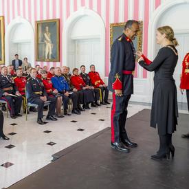 Satpal Singh Parhar receives his insignia from the Governor General.  RCMP Commissioner Brenda Lucki observes them standing behind them to the right.  All three are standing on stage in front of a room filled with people. 