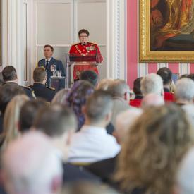 RCMP Commissioner Brenda Lucki stands on a stage in front of a podium facing a room filled with people.  She delivers a speech as the Governor General, seated on stage,  looks at her. 