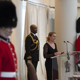 The Governor General stands in front of a podium and delivers a speech.  On either side of her are ceremonial guards.  Behind her, is her Aide-de-camps. 