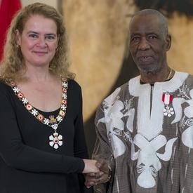 Lamine Touré se tient à côté de la Gouverneure générale.  Tous deux sourient à la caméra.  Ils portent leur insigne de l'Ordre du Canada.