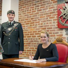 Governor General Julie Payette looks directly at the photographer, while she is sitting a table, pen in hand ready to sign a document. A men wearing a uniform is standing to her right.