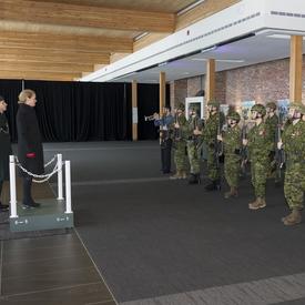 La gouverneure générale Julie Payette est debout sur un dais et regarde une garde de caserne composée de deux rangées de huit réservistes en uniforme de combat.
