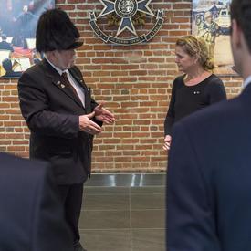 Un homme en complet et portant un chapeau à plumes noires s'adresse à la gouverneure générale qui l'écoute attentivement.