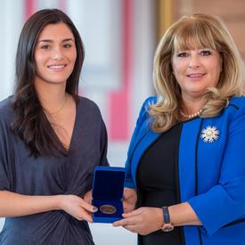 On the left, a female dark hair university student is holding an opened blue box containing a medal. A blond woman wearing a blue jacket is on the right.