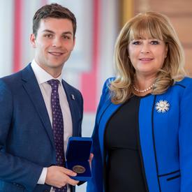 On the left, a male university student wearing a blue jacket is holding an opened blue box containing a medal. A blond woman wearing a blue jacket is on the right.