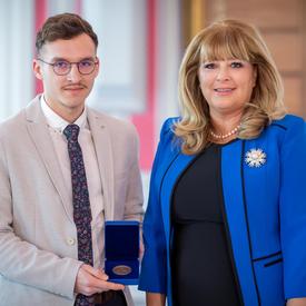 On the left, a male university student wearing a light gray jacket, is holding an opened blue box containing a medal. A blond woman wearing a blue jacket is on the right.