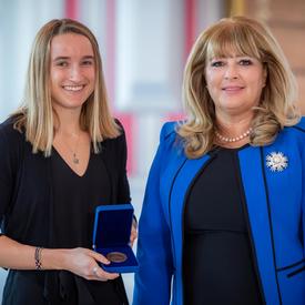 On the left, a blond female university student is holding an opened blue box containing a medal. A blond woman wearing a blue jacket is on the right.