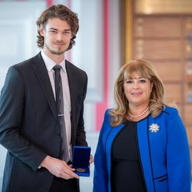 On the left, a tall male university student is holding an opened blue box containing a medal. A blond woman wearing a blue jacket is on the right.