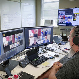 A man with a head set is sitting at a computer, looking at two large screens in front of him. A virtual conversation with 4 participants seems to be taking place.
