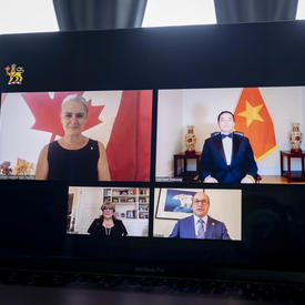 A laptop is opened in front of a large window with grey-blue draps. On the screen, we see 4 people, 2 women and 2 men, in 4 different rectangles. The flag of Vietnam in behind one of the men.