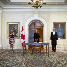 Five people standing in front of a pastel yellow wall. A table is in front of the person standing in the middle. A Canadian flag is placed to her right.