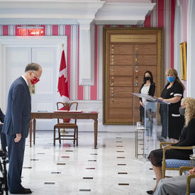 Un homme en costume est debout devant la gouverneure générale qui est assis. Il la reconnaît d'un signe de tête. Le Premier ministre regarde sur le côté.
