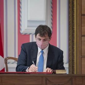 Un homme est assis à un bureau à la droite d'un grand drapeau canadien. Il est en train de signer un document.