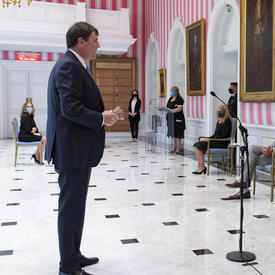 Deux hommes sont debout, l'un en face de l'autre. L'homme de droite tient un document que l'autre homme peut voir. L'homme qui lit le document parle dans un microphone.
