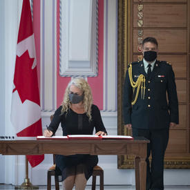 A woman is seated at a desk between a large Canadian flag and a soldier in dress uniform. She is signing a document.