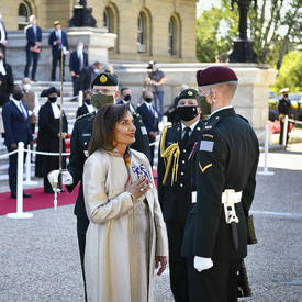 A woman dressed in white stands in front of a soldier in service dress. Hius uniform has one chevron on the left arm. The woman has her right hand pressed to her chest and she is smiling.