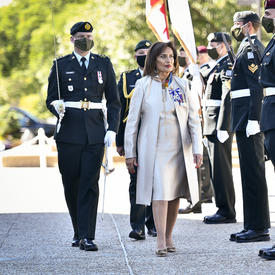 A woman dressed in white is accompanied by a military member in service dress carrying a sword. They are walking past a straight line of military members stood at attention.