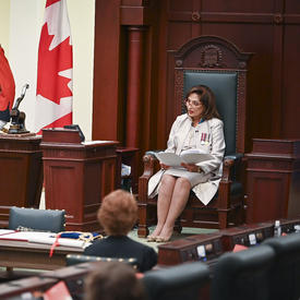 A woman dressed in white sits on a throne made of dark wood and green leather. She is reading from a document. To her right are a large Canadian flag and two military members on service dress.