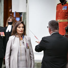 A man in a dark suit touches the shoulder of a woman dressed in white with a metal sword. Standing behind the woman are a military member in service dress and three people dressed in black ceremonial attire. One is holding a black staff with metal trim.