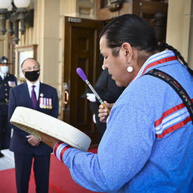 A man dressed in a light blue jacket with red trim plays a small drum held in his left hand. A woman dressed in white and a man in a dark suit are watching.