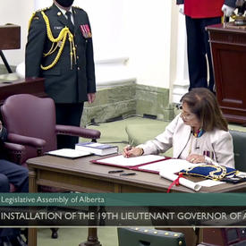 A woman dressed in white sits at a table. She is signing a document. Looking on are a man in a dark suit and a military member in service dress.